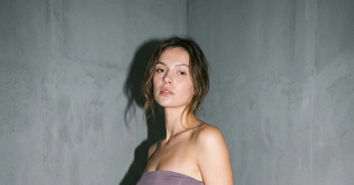 Woman with brown hair pulled back standing against a concrete wall and looking at the camera