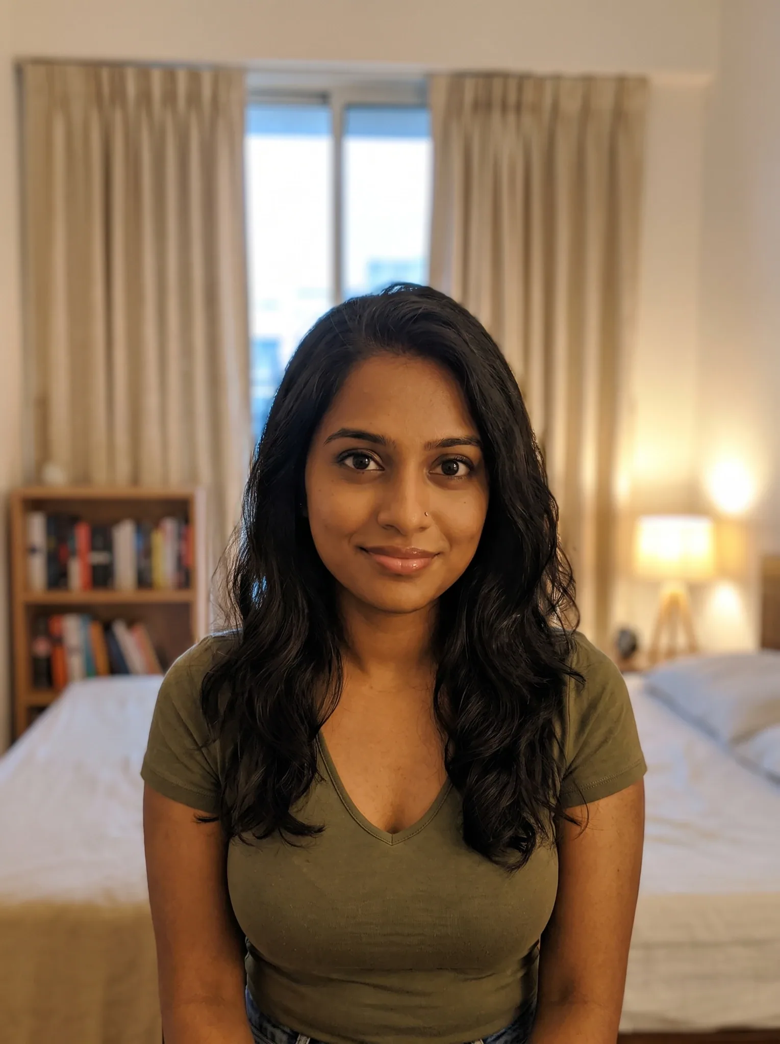 Priya, an adult AI character, looking at the camera in a warmly lit bedroom, wearing an olive T-shirt, with long wavy dark hair