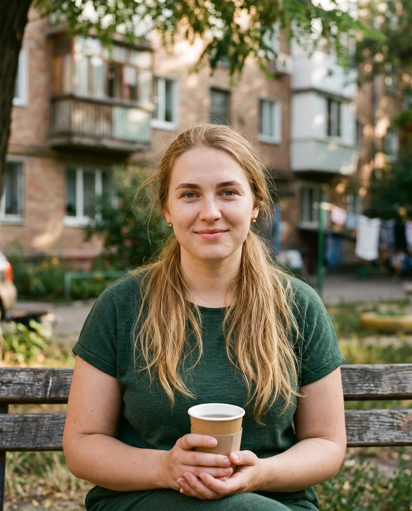 Zlata, an adult AI character, sitting on a park bench holding a paper coffee cup, with a brick apartment block behind her