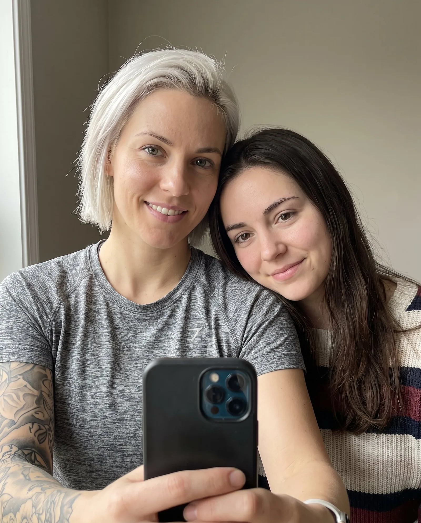Kira and Lou, the couple character, taking a mirror selfie together: one has short platinum hair and a tattooed arm, the other long dark hair