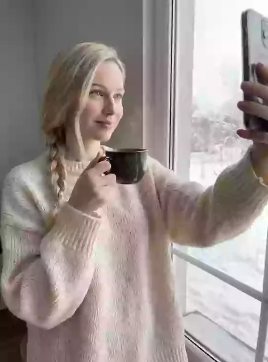 Lena, an adult AI character, holding a coffee mug and taking a selfie in a cream knit sweater